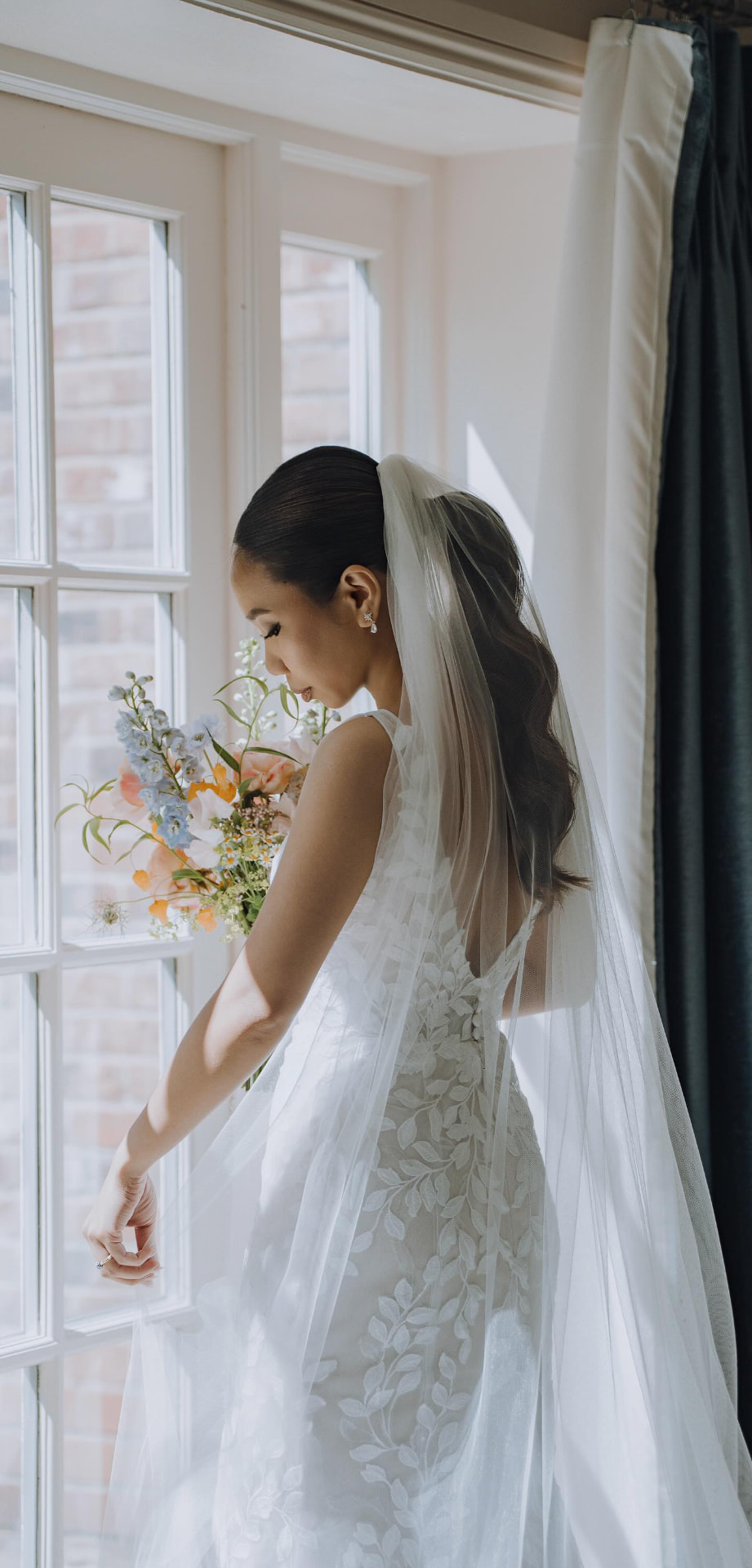 Profile of a bride with a long ponytail and veil, standing beside a window.