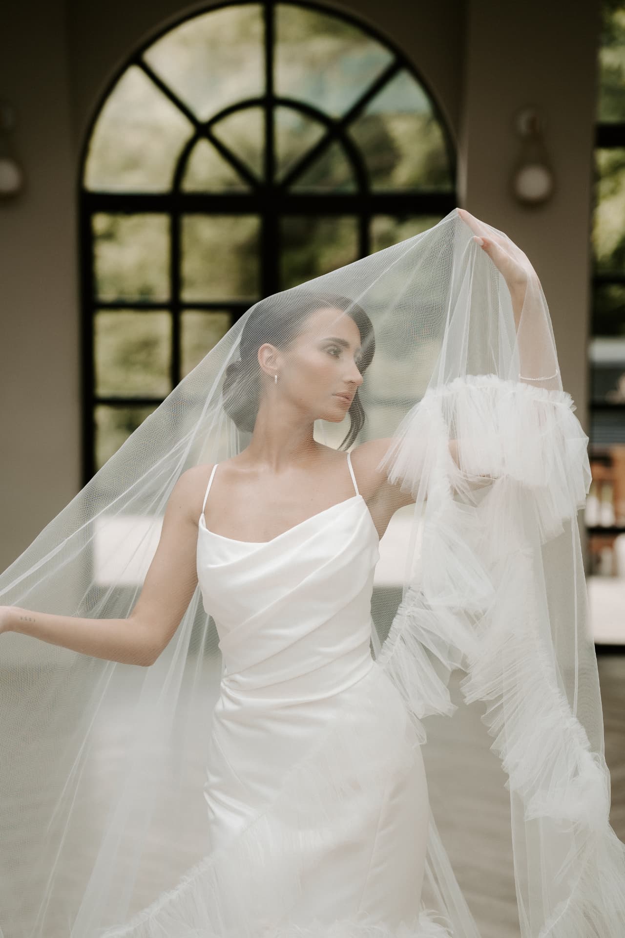 Bride holding a ruffled veil above her head in front of an arched window.