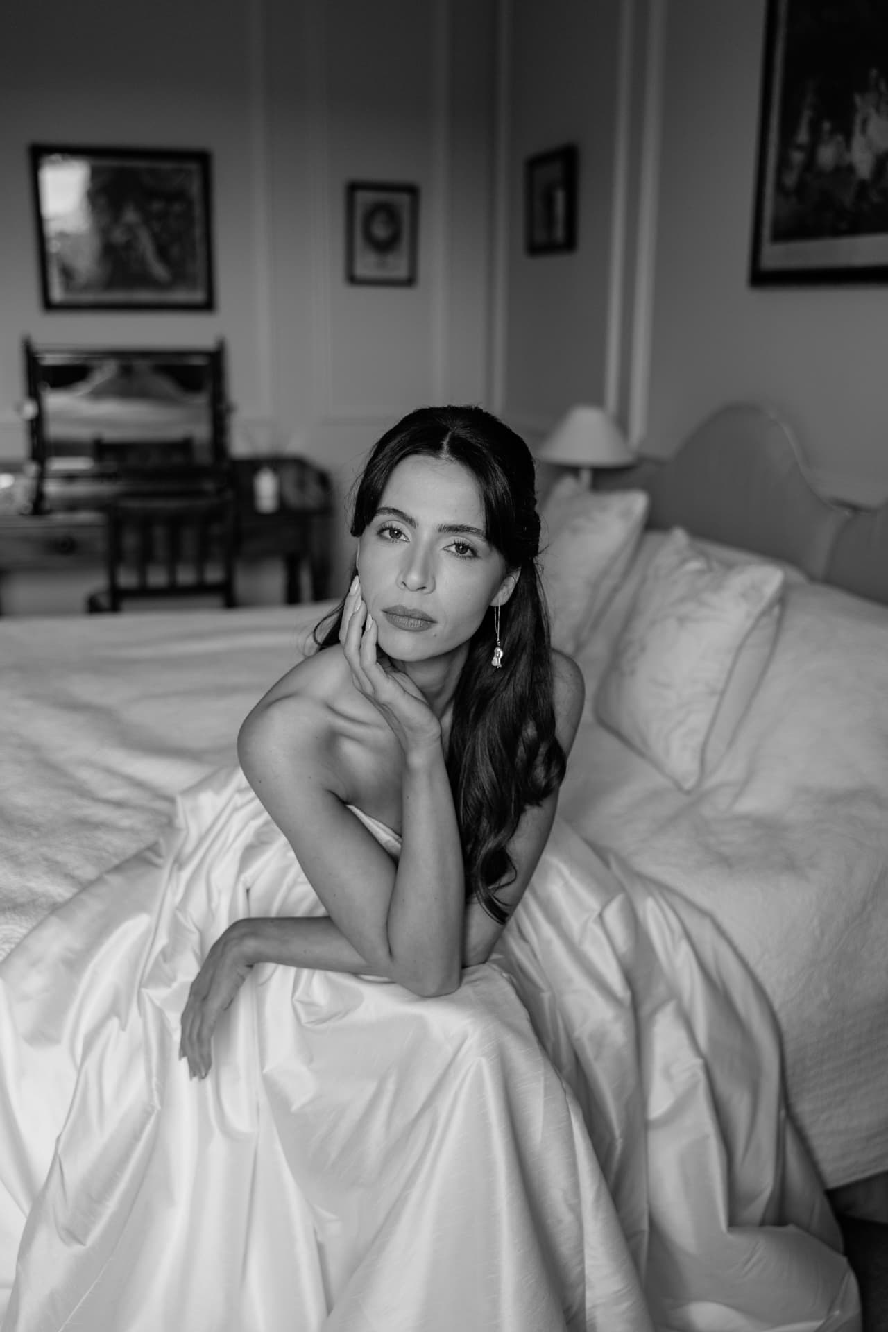 Black and white portrait of a bride seated on a bed in a full taffeta skirt.