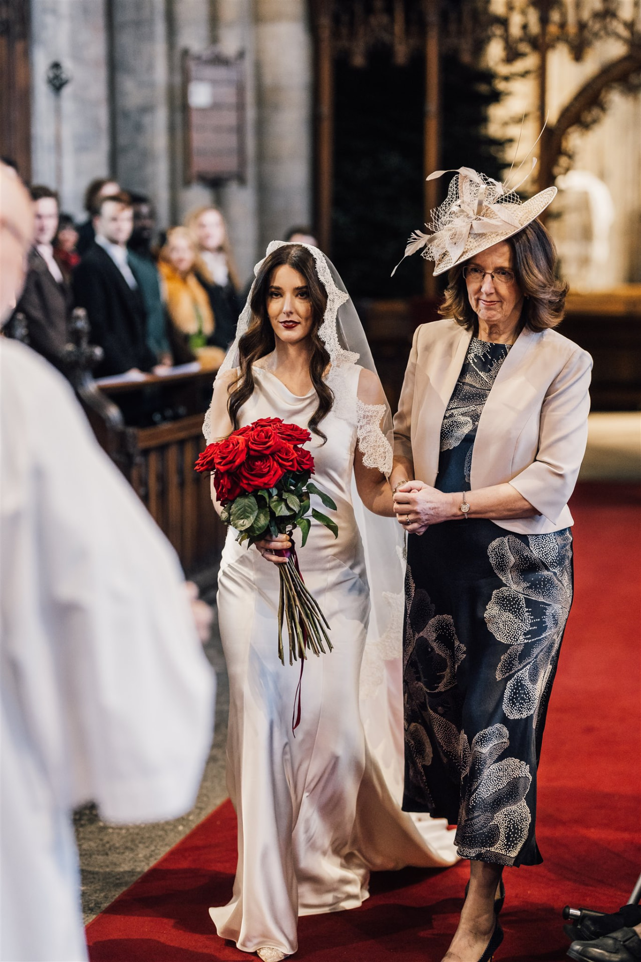 Bride with long brown waves and red roses walking down the aisle with her mother.