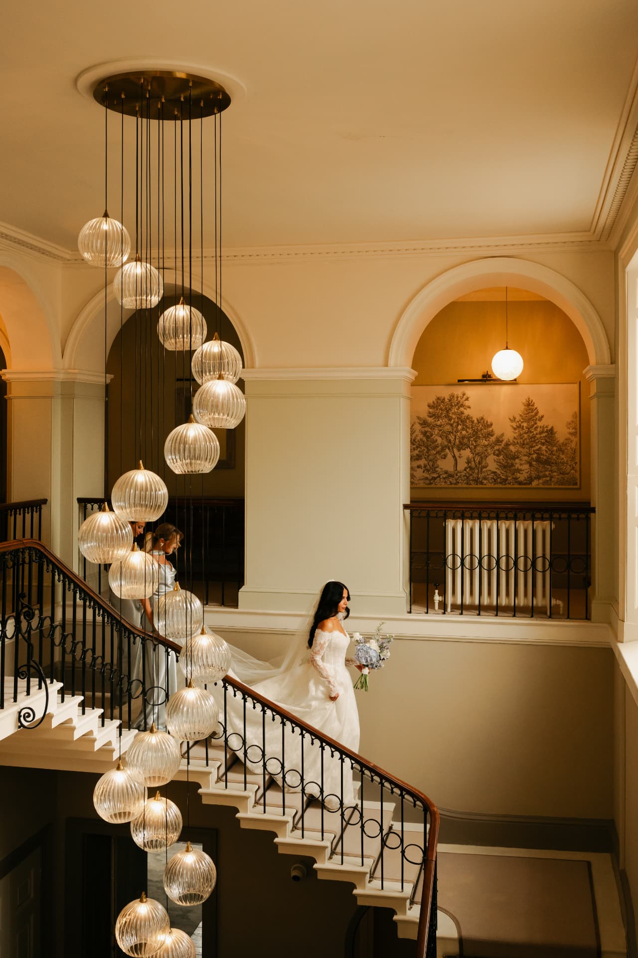 Bride descending a sweeping country-house staircase beneath a cascade of glass pendant lights.