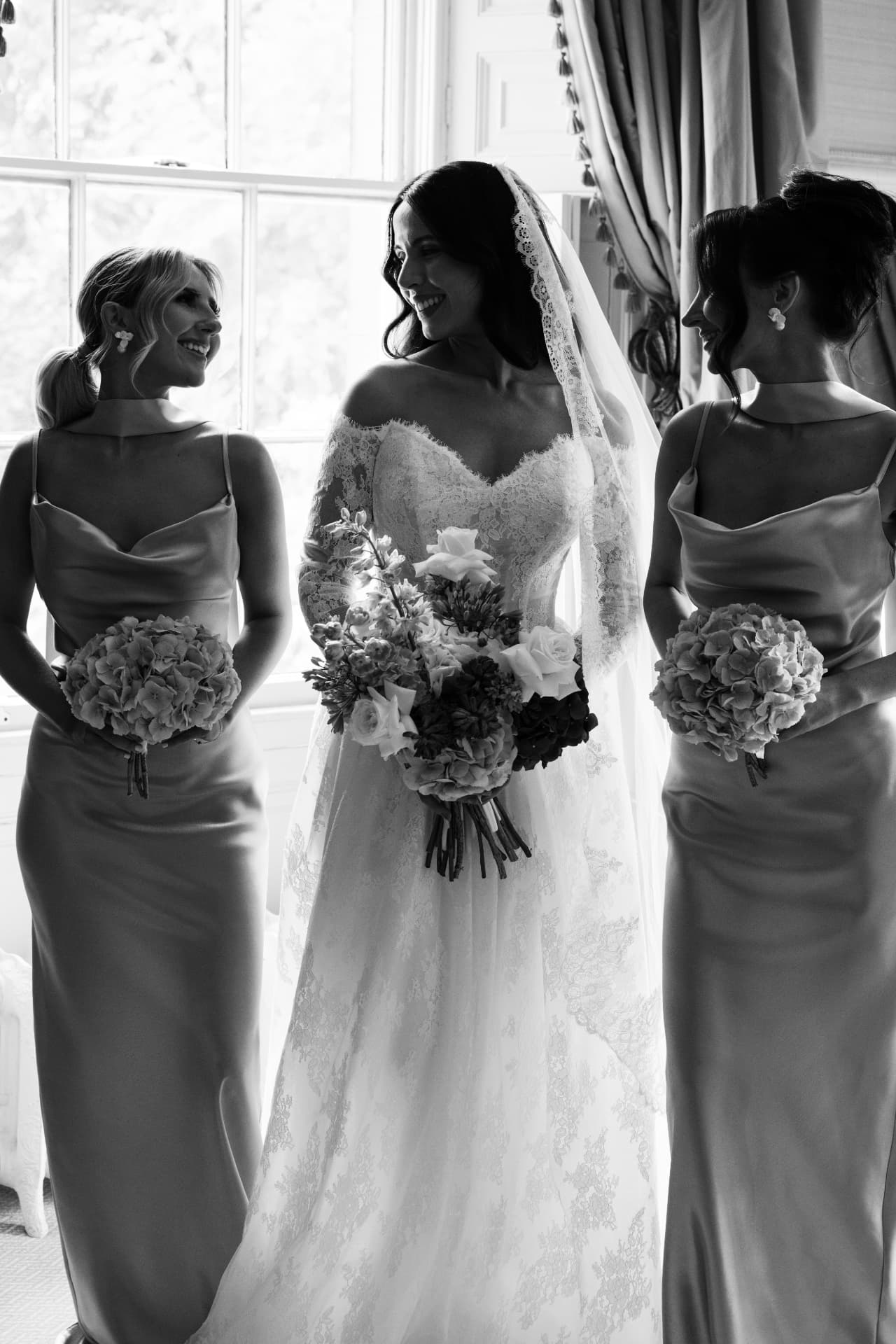 Black and white photograph of a bride laughing with her two bridesmaids at a window.