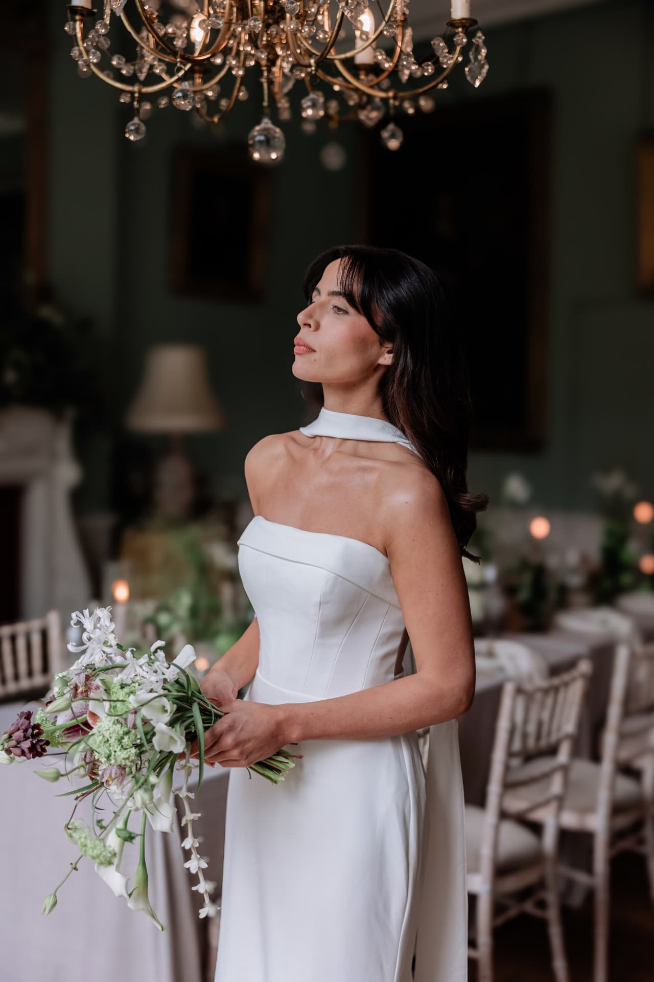Bride standing beneath a crystal chandelier holding a trailing bouquet.
