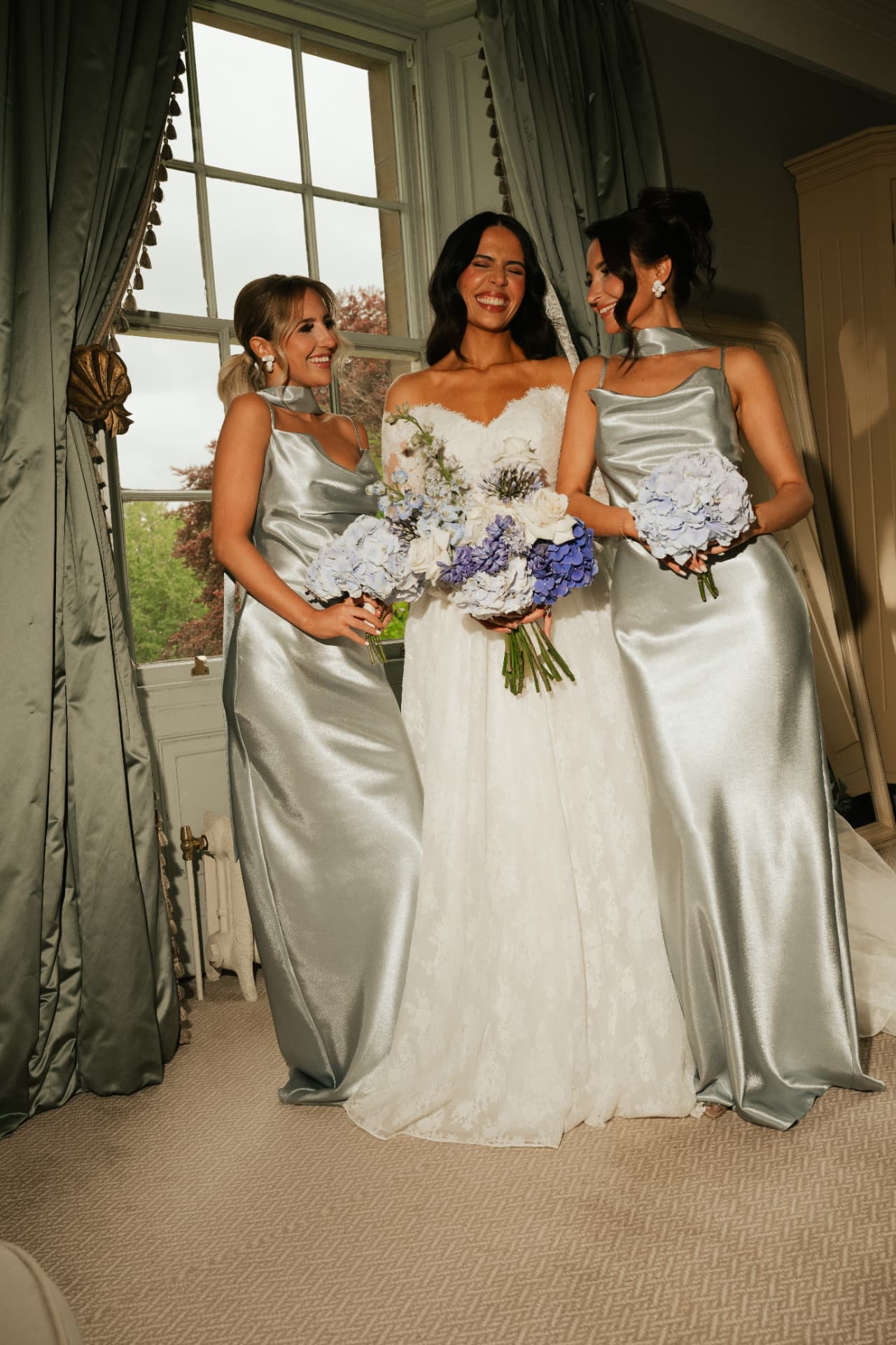 Bride laughing between two bridesmaids in silver satin, holding hydrangeas.